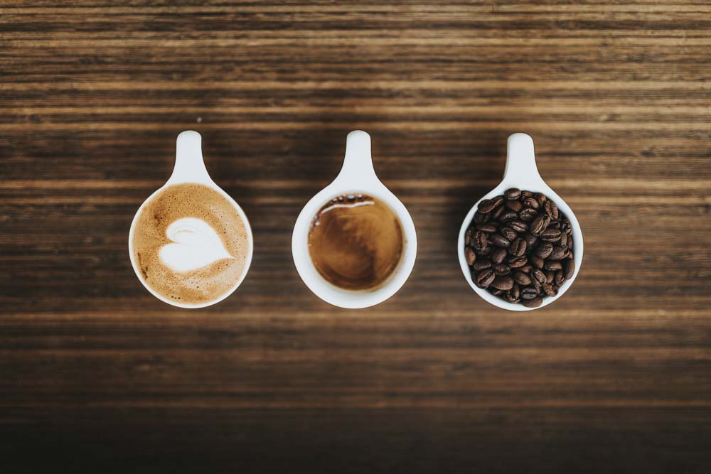 Three ceramic cups. One contains coffee beans, one espresso, another has latte art with a heart